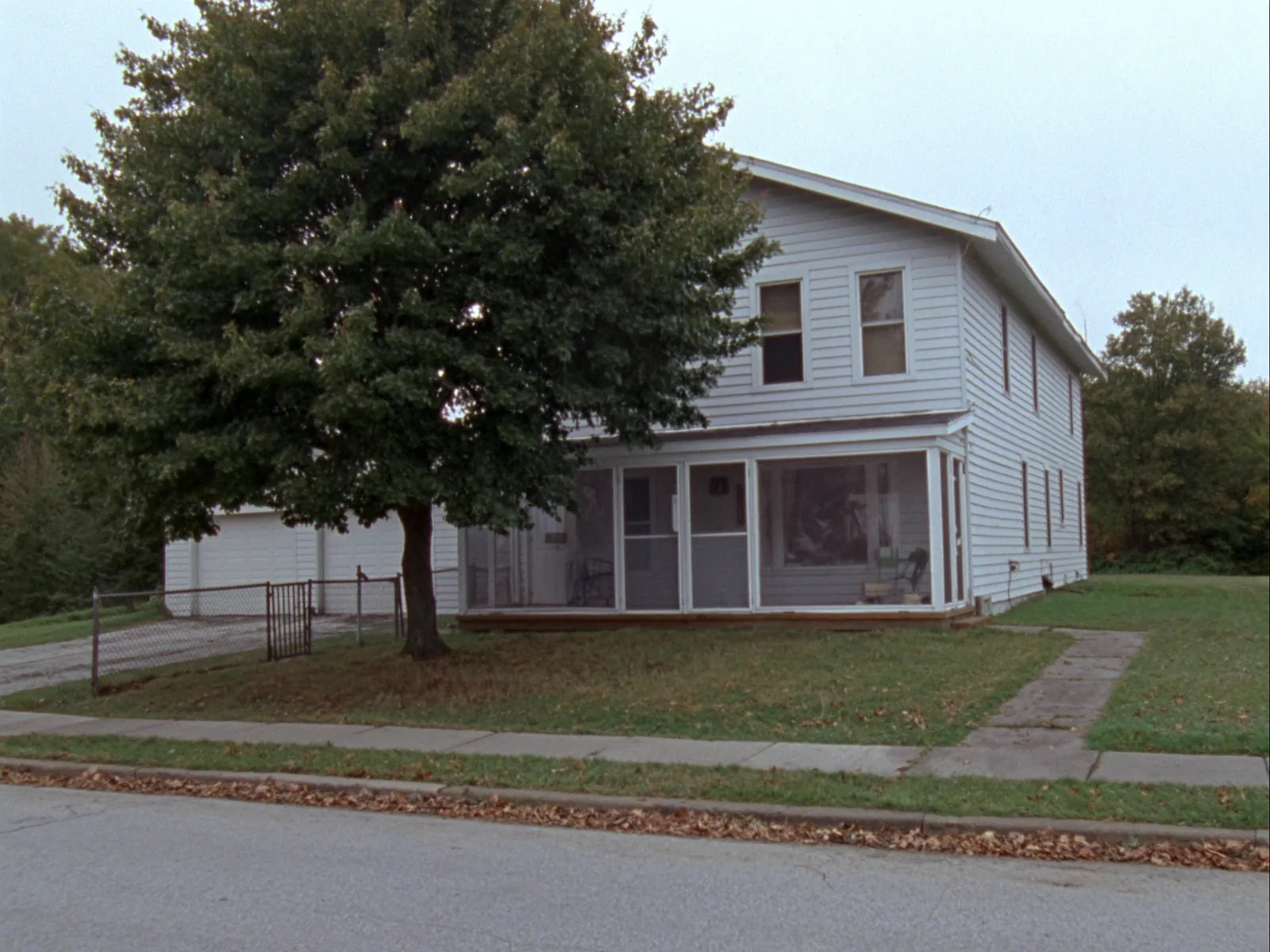 A white two-story frame house with a screened front porch, a large maple in the yard, a chain-link fence and a detached garage down a gravel drive on a small-town street.