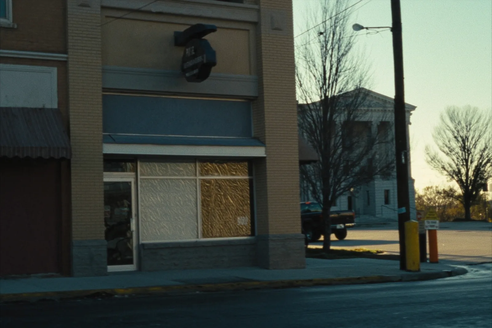 A small brick storefront office with a plate glass window on a courthouse square, a pickup truck parked at an angle at the curb, bare winter trees and a limestone courthouse across the street.