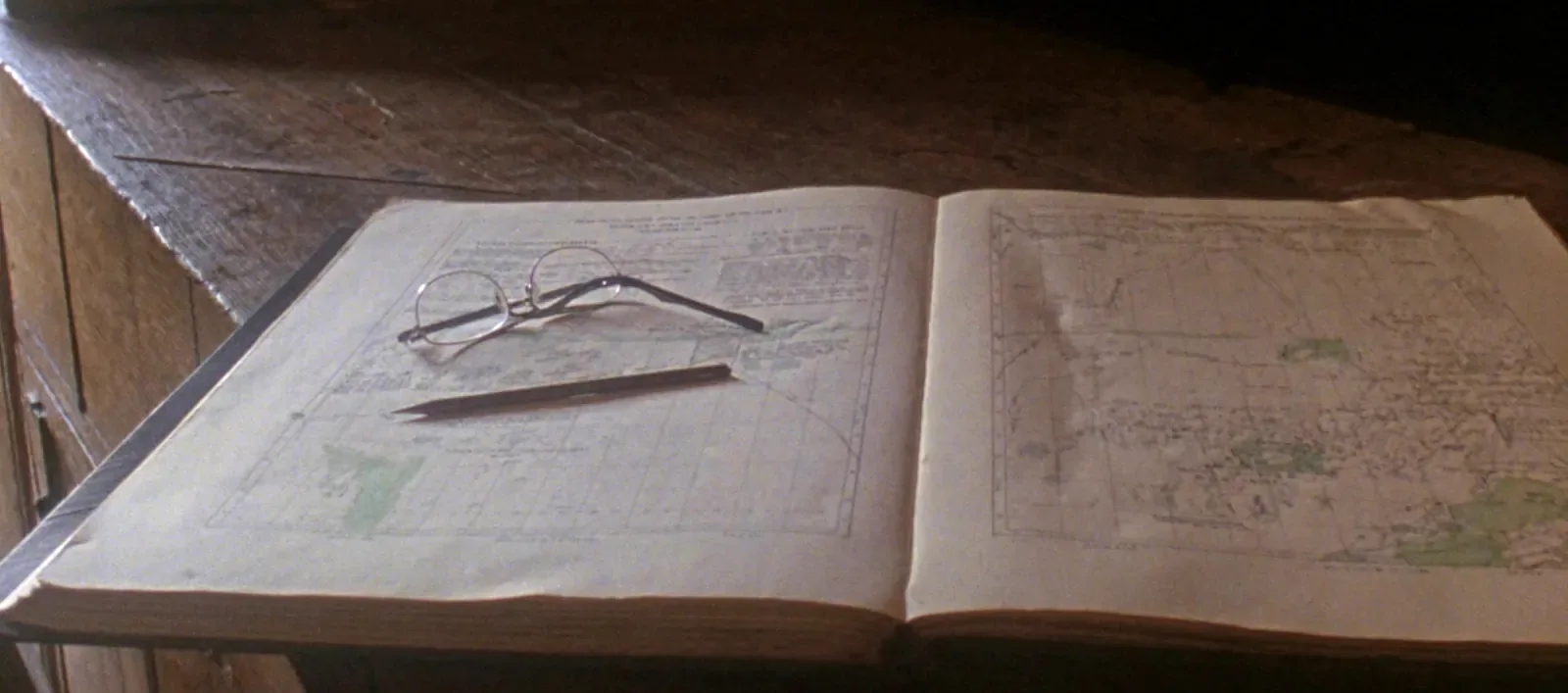 An open plat book on a worn wooden counter showing two pages of surveyed land divided into a fine grid, with reading glasses and a pencil resting on the left page and stacked older volumes behind.