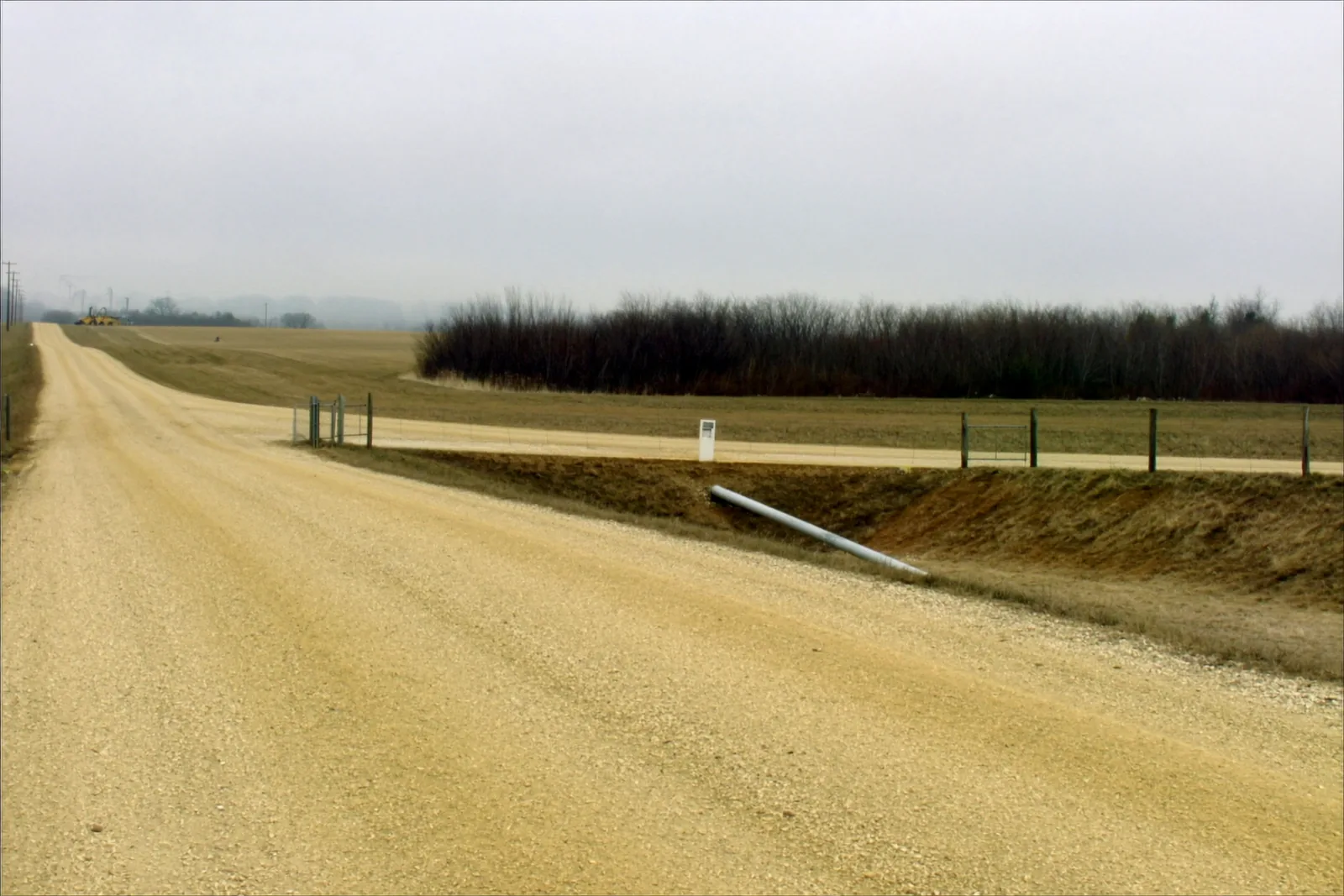 A gravel county road running between open fields under a grey sky, with a ditch and a wire fence along the far side, a metal gate at a field entrance and a length of culvert pipe lying in the ditch.